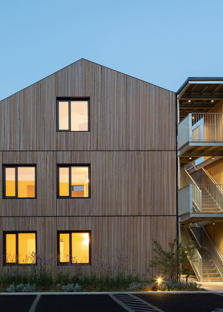 Modern timber-clad residential building at dusk with warm interior lighting visible through large windows, surrounded by landscaped gardens